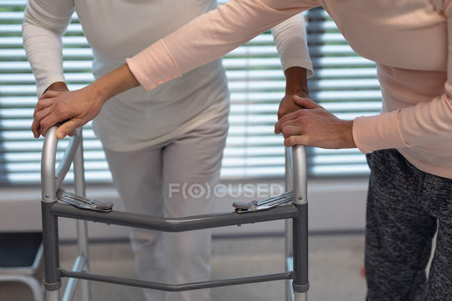 Низька секція жіночого фізіотерапевта, який допомагає пацієнтці змішаної раси ходити з ходунками в лікарні — стокове фото