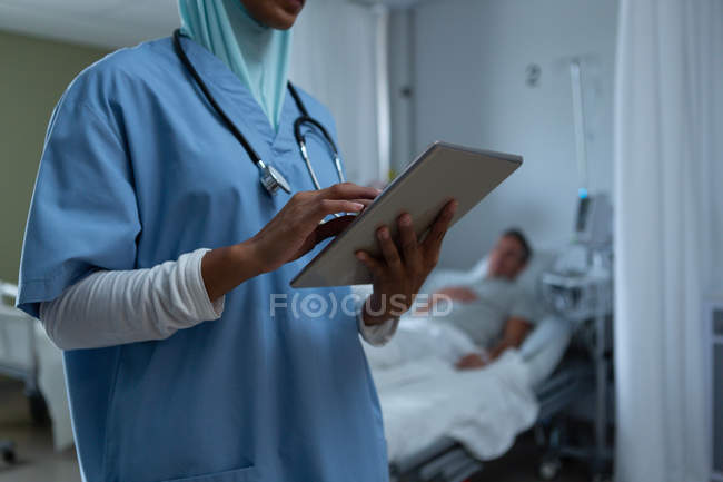 Seção intermediária de médico feminino de raça bastante mista em hijab usando comprimido digital na enfermaria enquanto paciente masculino caucasiano dorme em segundo plano no hospital — Fotografia de Stock