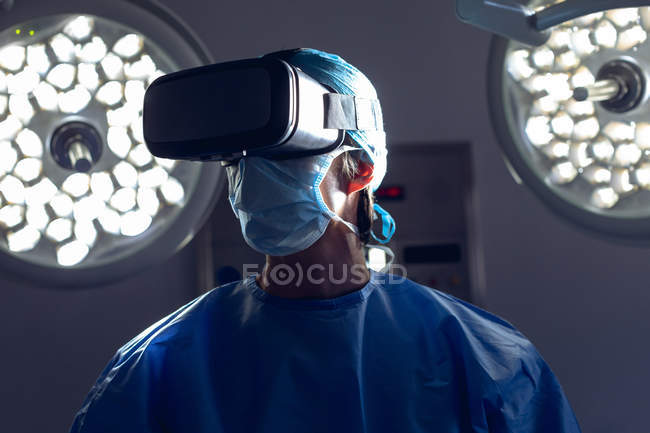 Visão frontal de cirurgiã branca madura usando fone de ouvido de realidade virtual na sala de cirurgia do hospital. Luzes médicas e monitores em segundo plano . — Fotografia de Stock