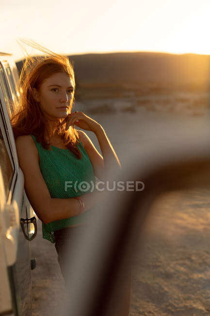 Front view of beautiful young Caucasian woman leaning on camper van at beach — Stock Photo