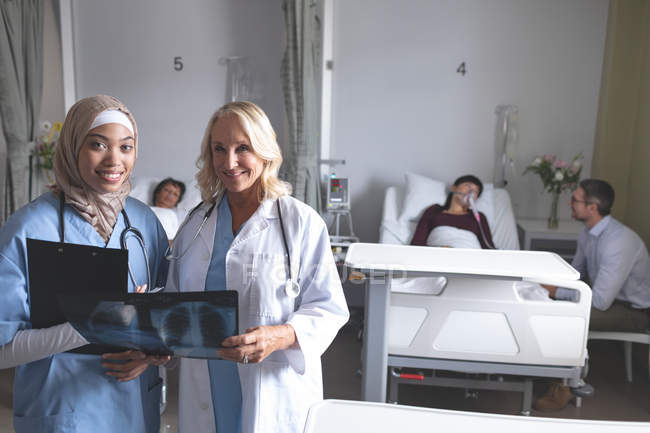 Front view of diverse female doctors discussing over x-ray report in the ward at hospital. In the background diverse patients are lying in their beds. — Stock Photo