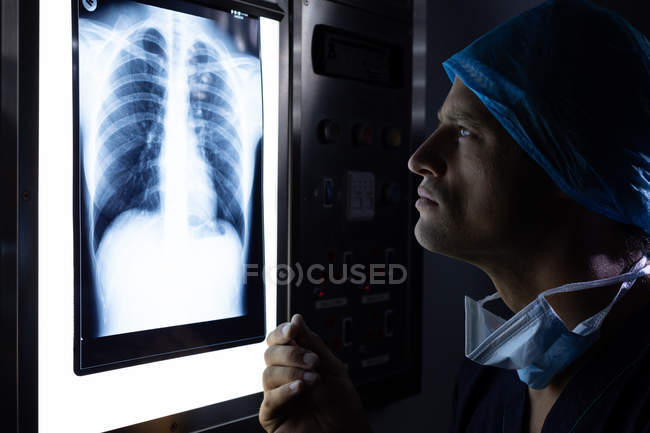 Vista lateral del guapo joven cirujano caucásico examinando rayos X en caja de luz en quirófano en el hospital . - foto de stock