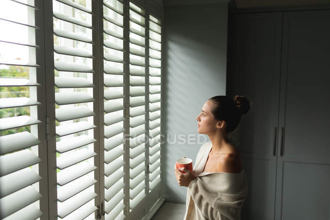 Vista lateral de mujer caucásica con taza de café mirando a través de la ventana en casa - foto de stock