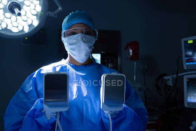 Retrato de una joven cirujana de raza mixta que sostiene el desfibrilador en el quirófano del hospital . - foto de stock