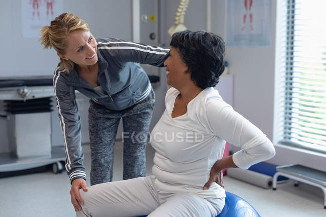 Vue latérale du physiothérapeute féminin caucasien aidant la patiente métisse sur le ballon d'exercice à l'hôpital — Photo de stock