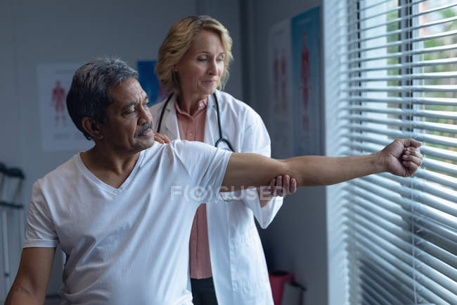 Vista frontal del médico femenino caucásico maduro examinando el brazo de paciente masculino de raza mixta senior en el hospital - foto de stock