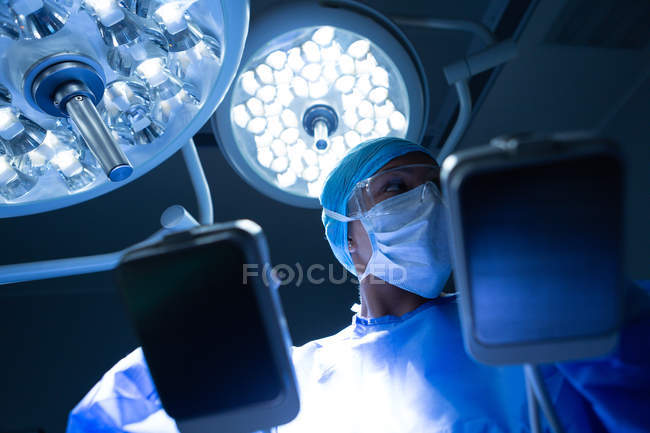 Visão de baixo ângulo de cirurgião feminino de raça mista segurando desfibrilador enquanto olha para longe no teatro de operação no hospital — Fotografia de Stock