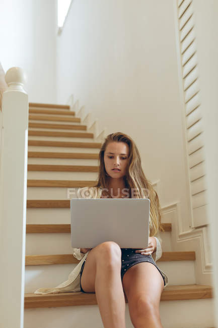 Vue de face de belle femme caucasienne en utilisant un ordinateur portable sur les escaliers à la maison — Photo de stock