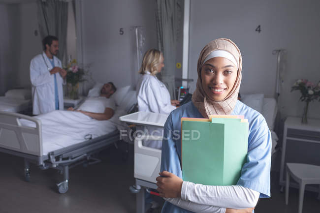 Vista frontal de uma médica mestiça em hijab, com arquivos médicos na enfermaria do hospital. No fundo, médicos caucasianos verificando os pacientes . — Fotografia de Stock