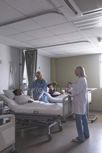 Vista lateral de diversos médicos que interactúan con pacientes en la sala del hospital. En el fondo el hombre caucásico está sosteniendo la mano de la mujer asiática que está acostada en la cama en el hospital . - foto de stock