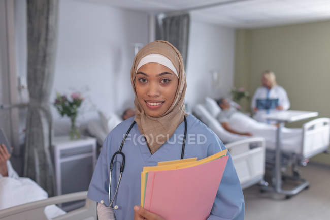 Портрет женщины-врача смешанной расы в хиджабе, стоящей с медицинскими документами в палате больницы. На заднем плане различные пациенты, лежащие в постели и кавказская женщина врач проверяет их . — стоковое фото