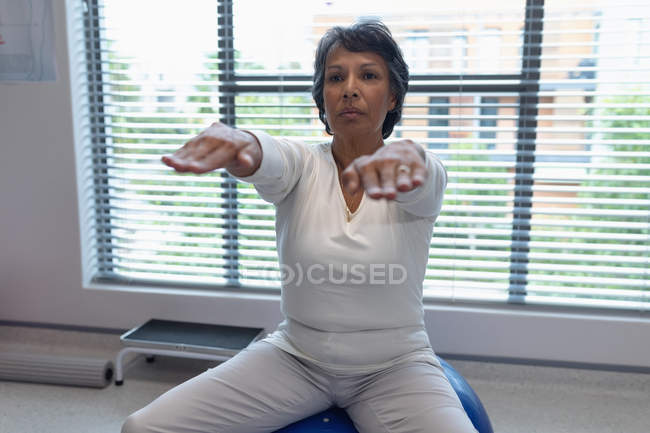 Vista frontal de una paciente de raza mixta que hace ejercicio sobre una pelota de ejercicio en el hospital - foto de stock