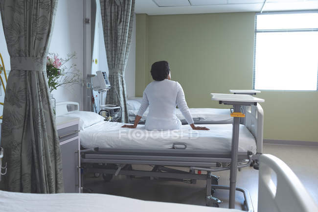 Rear view of mixed race female patient sitting on bed in the ward at hospital — Stock Photo