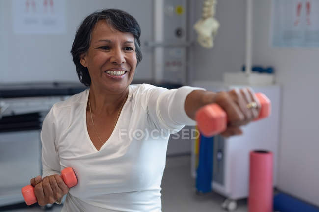 Vista frontal de una paciente de raza mixta que hace ejercicio con pesas anaranjadas en el hospital - foto de stock