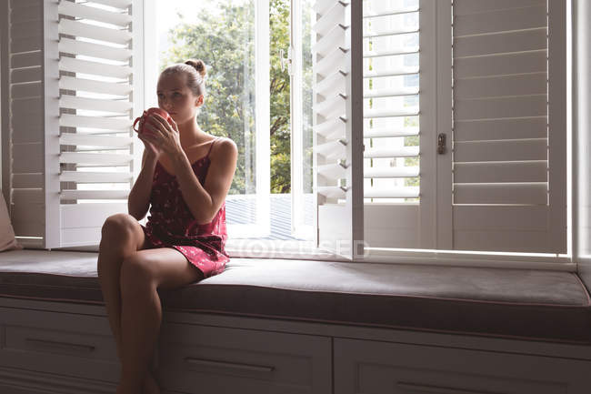 Vista frontale della bella donna caucasica che beve caffè mentre è seduta sul sedile del finestrino in camera da letto a casa — Foto stock