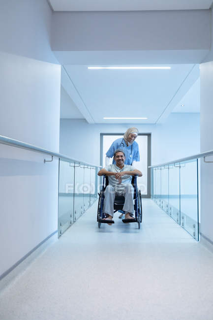 Вид спереду жінка-лікар змішаної раси штовхає старшого пацієнта змішаної раси в інвалідному кріслі в коридорі в лікарні . — стокове фото
