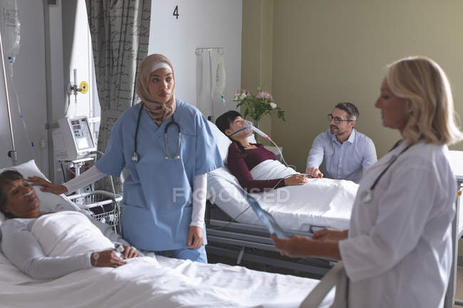 Vue de face de divers médecins féminins qui interagissent avec des patientes à l'hôpital. Dans le fond l'homme caucasien tient la main de la femme asiatique qui est couchée dans le lit à l'hôpital . — Photo de stock