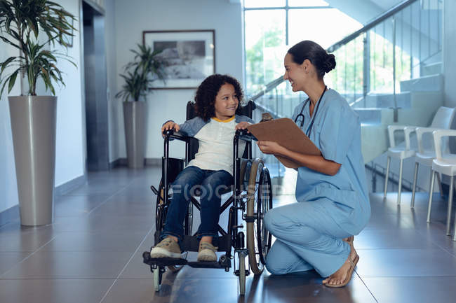 Vista lateral de la feliz doctora caucásica interactuando con un niño discapacitado de raza mixta en el pasillo del hospital. Chico está sentado en silla de ruedas . - foto de stock