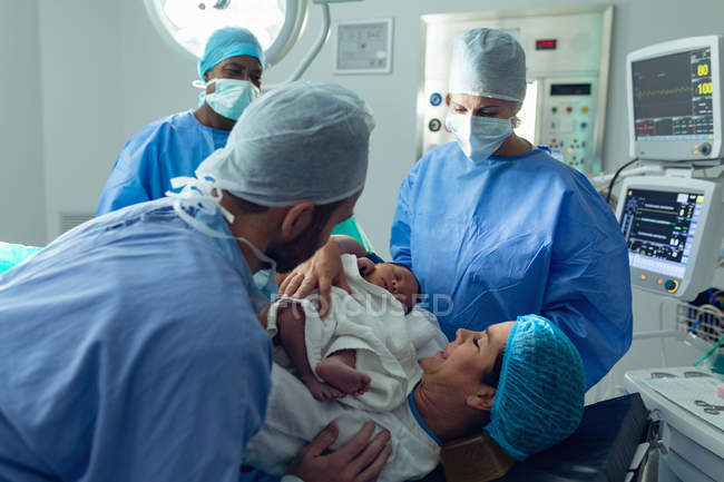 Vista lateral de pareja caucásica con cirujanos sosteniendo a su bebé recién nacido en quirófano en el hospital - foto de stock