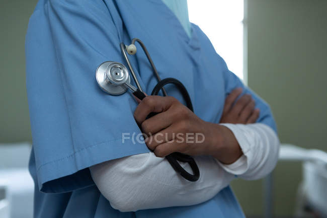Средняя часть женщины-врача, стоящей со скрещенными руками, держа стетоскоп в руке в палате больницы — стоковое фото