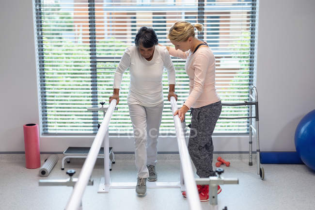 Vue de face du physiothérapeute caucasien aidant une patiente métisse à marcher avec des barres parallèles à l'hôpital — Photo de stock