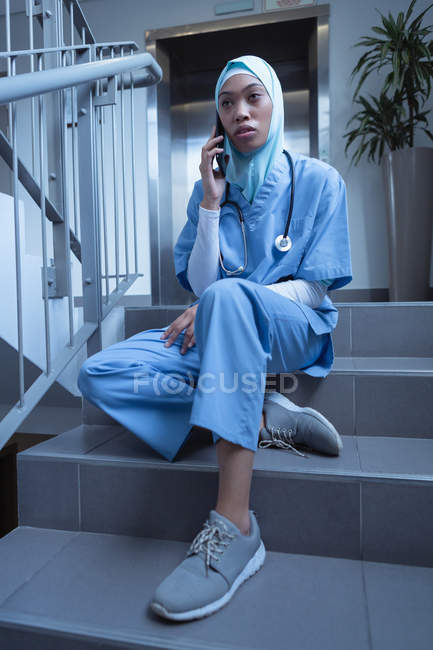 Вид спереду змішаної раси жінка медсестра в хіджабі розмовляє на мобільному телефоні, сидячи на сходах в лікарні — стокове фото