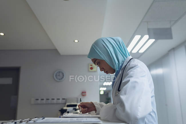 Vista laterale del medico di razza mista nei documenti di lettura hijab alla reception in ospedale — Foto stock