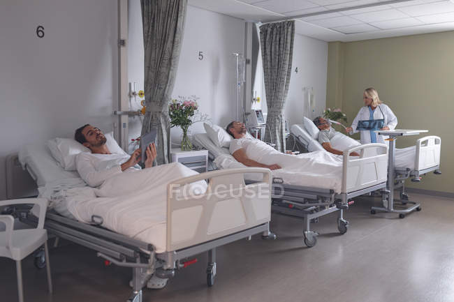 Visão geral de diversos pacientes do sexo masculino usando comprimido digital ou dormindo enquanto o médico feminino verifica o paciente masculino na enfermaria do hospital — Fotografia de Stock