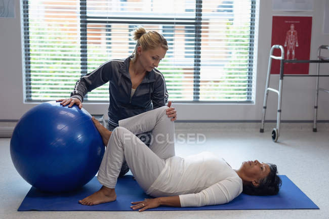 Vista lateral de fisioterapeuta femenina caucásica ayudando a paciente de raza mixta con ejercicio sobre pelota de ejercicio en el hospital . - foto de stock