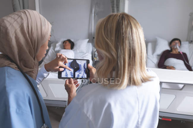 Rear view of diverse female doctors discussing over x-ray report on digital tablet in the ward at hospital. Diverse female patients are sleeping in bed in the background. — Stock Photo