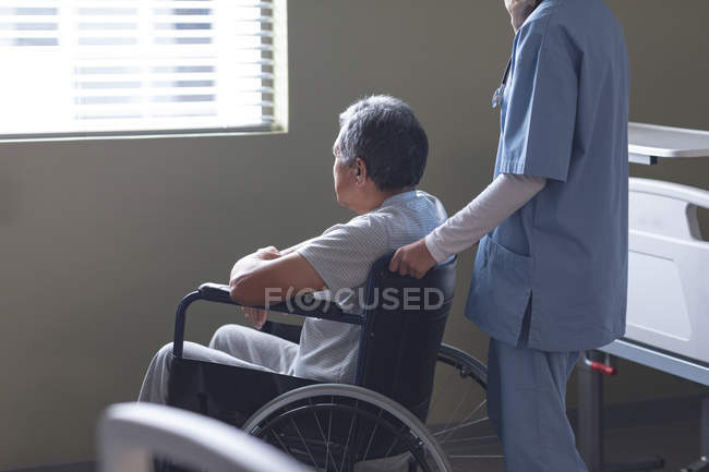 Side view of mixed-race female doctor standing with senior mixed-race male patient in wheelchair at hospital — Stock Photo