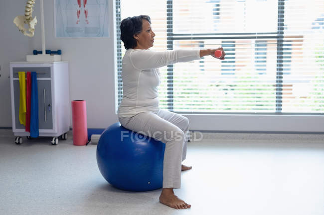 Vista lateral de una paciente de raza mixta que hace ejercicio con pesas sobre una pelota de ejercicio en el hospital - foto de stock