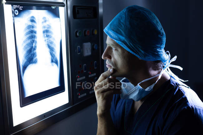 Vista laterale del bel giovane chirurgo maschio caucasico che esamina i raggi X sulla scatola luminosa in sala operatoria in ospedale . — Foto stock