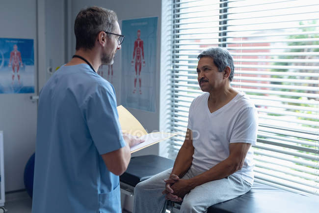 Side view of handsome Caucasian male surgeon interacting with senior mixed-race patient while holding medical files in his hands in hospital — Stock Photo