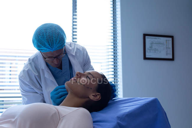 Vorderseite des hübschen kaukasischen Chirurgen mittleren Alters, der das schöne Gesicht der kaukasischen Frau vor der Operation im Krankenhaus markiert — Stockfoto