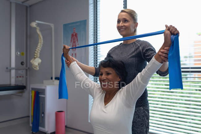 Vista lateral de fisioterapeuta femenina caucásica que da fisioterapia con banda de resistencia a paciente femenina de raza mixta en el hospital - foto de stock