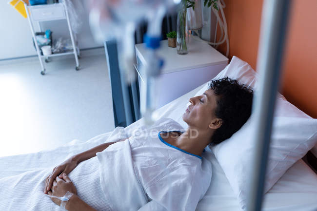 Side view of middle aged mixed-race female patient sleeping in bed with hands on stomach in the ward at hospital — Stock Photo