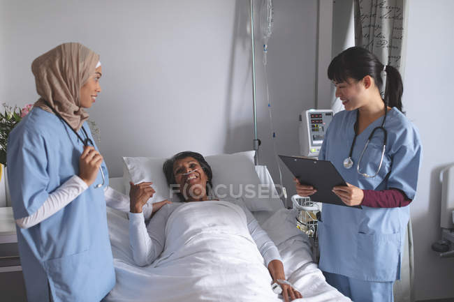 Vista frontal de diversas doctoras que interactúan con una paciente madura de raza mixta en la sala del hospital. Paciente femenina se ve feliz . - foto de stock