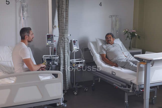 Visão lateral de diversos pacientes do sexo masculino interagindo uns com os outros enquanto relaxa na cama na enfermaria do hospital . — Fotografia de Stock