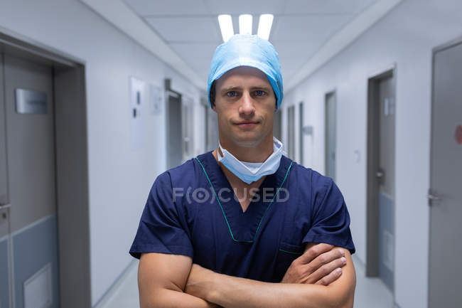 Retrato de um belo jovem cirurgião caucasiano de pé com os braços cruzados no corredor do hospital. O homem está usando máscara cirúrgica, vestido e boné . — Fotografia de Stock