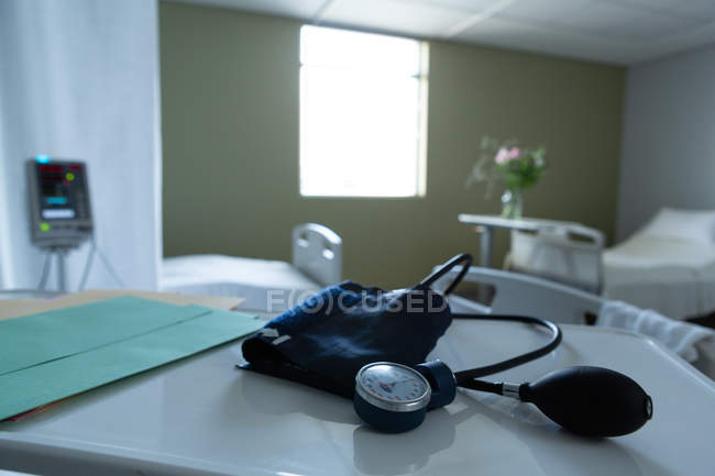 Крупный план измерителя артериального давления и медицинских карт на столе с пустыми кроватями и монитором на заднем плане в палате больницы — стоковое фото