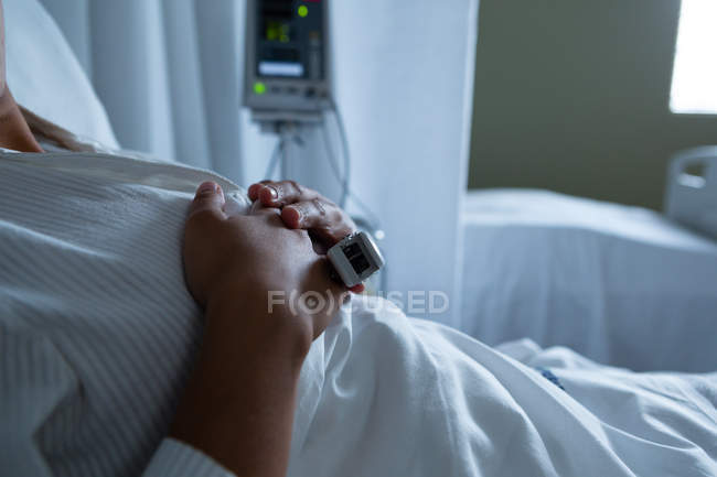 Mittelteil einer Patientin, die im Bett auf der Station liegt, mit den Händen auf ihren Brüsten im Krankenhaus. Im Hintergrund sind leeres Bett und Monitor zu sehen. — Stockfoto