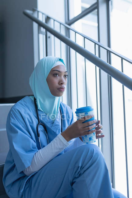Side view of thoughtful mixed race female doctor in hijab having coffee on staircase in hospital — Stock Photo