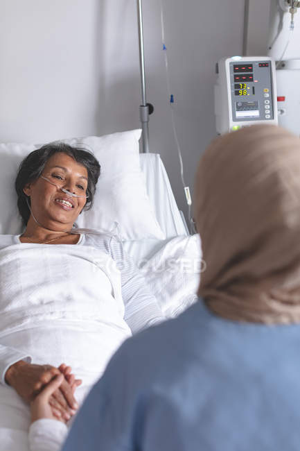 Над видом на плече змішаної раси жінка-лікар в хіджабі втішає красивого пацієнта змішаної раси в палаті в лікарні. Вони тримають руки . — стокове фото