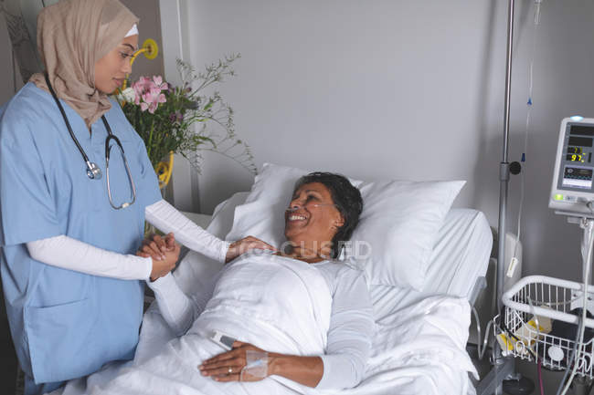 Visão frontal da mestiça médica feminina no hijab consolando a mestiça madura paciente feminina na enfermaria do hospital . — Fotografia de Stock