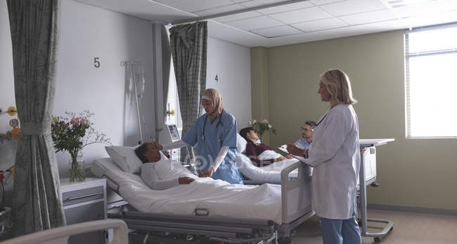 Visão lateral de diversos médicos interagindo com pacientes na enfermaria do hospital. No fundo homem caucasiano está segurando a mão da mulher asiática que está deitada na cama no hospital . — Fotografia de Stock