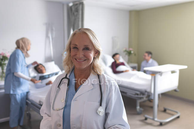 Portrait of Caucasian female doctor smiling in the ward at hospital. In the background diverse doctors are interacting with their patients next to the beds. — Stock Photo