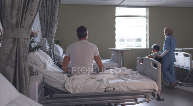Vista trasera del paciente varón caucásico sentado en la cama y la mujer de raza mixta médico en hiyab con discapacidad sénior de raza mixta paciente masculino en la sala en el hospital - foto de stock