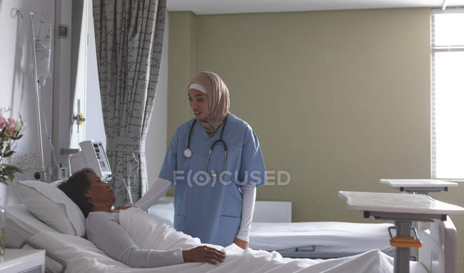 Vista lateral de una hermosa enfermera de raza mixta en hijab interactuando con una paciente de raza mixta en la sala del hospital. La enfermera tiene estetoscopio alrededor del cuello. . - foto de stock