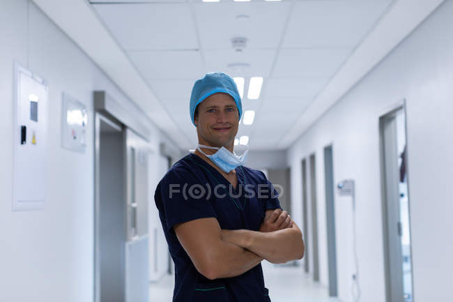 Portrait d'un beau jeune chirurgien caucasien debout les bras croisés dans le couloir de l'hôpital. L'homme porte un masque chirurgical, une robe et un bonnet . — Photo de stock